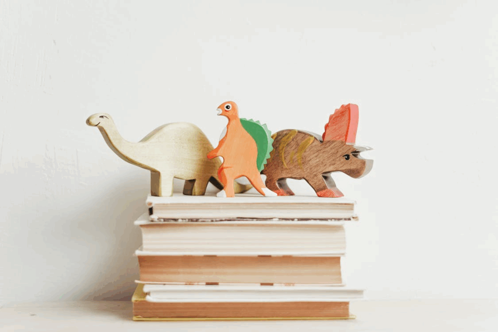 三個不同顏色和形狀的木製恐龍玩具（包括一隻長頸恐龍），疊放在一堆整齊的棕色書本上，象徵著童年學習、想像力或教育