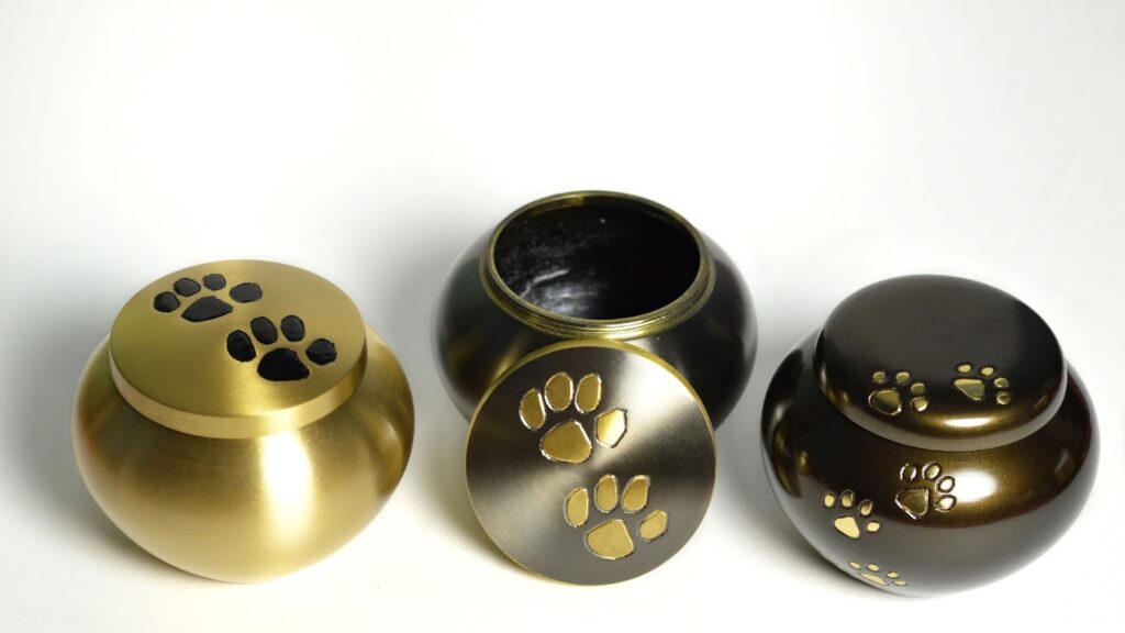 三個不同色調的圓形金屬寵物骨灰罐,分別為金色、黑色和古銅色,裝飾有精緻的金色爪印圖案
