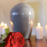 木質桌面上一朵盛開的紅玫瑰與數支點燃的白色蠟燭，背景為模糊的灰色骨灰罈。