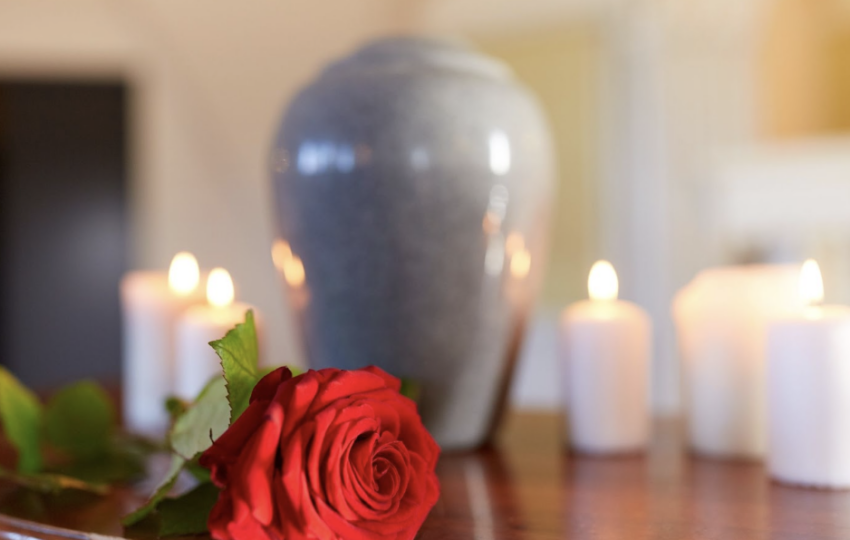 木質桌面上一朵盛開的紅玫瑰與數支點燃的白色蠟燭，背景為模糊的灰色骨灰罈。