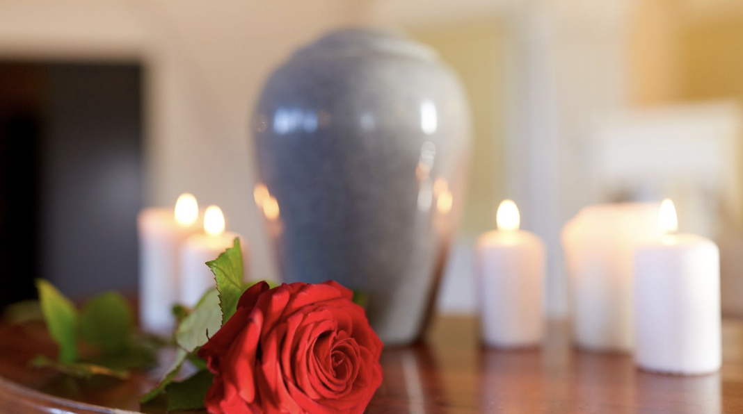 木質桌面上一朵盛開的紅玫瑰與數支點燃的白色蠟燭,背景為模糊的灰色骨灰罈。